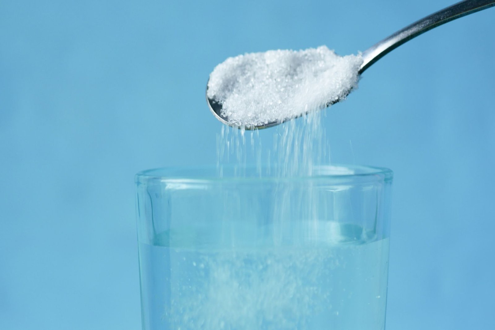 pouring of white sugar from spoon in a clear drinking glass with water