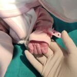 newborn in pink outfit holding gloved hand