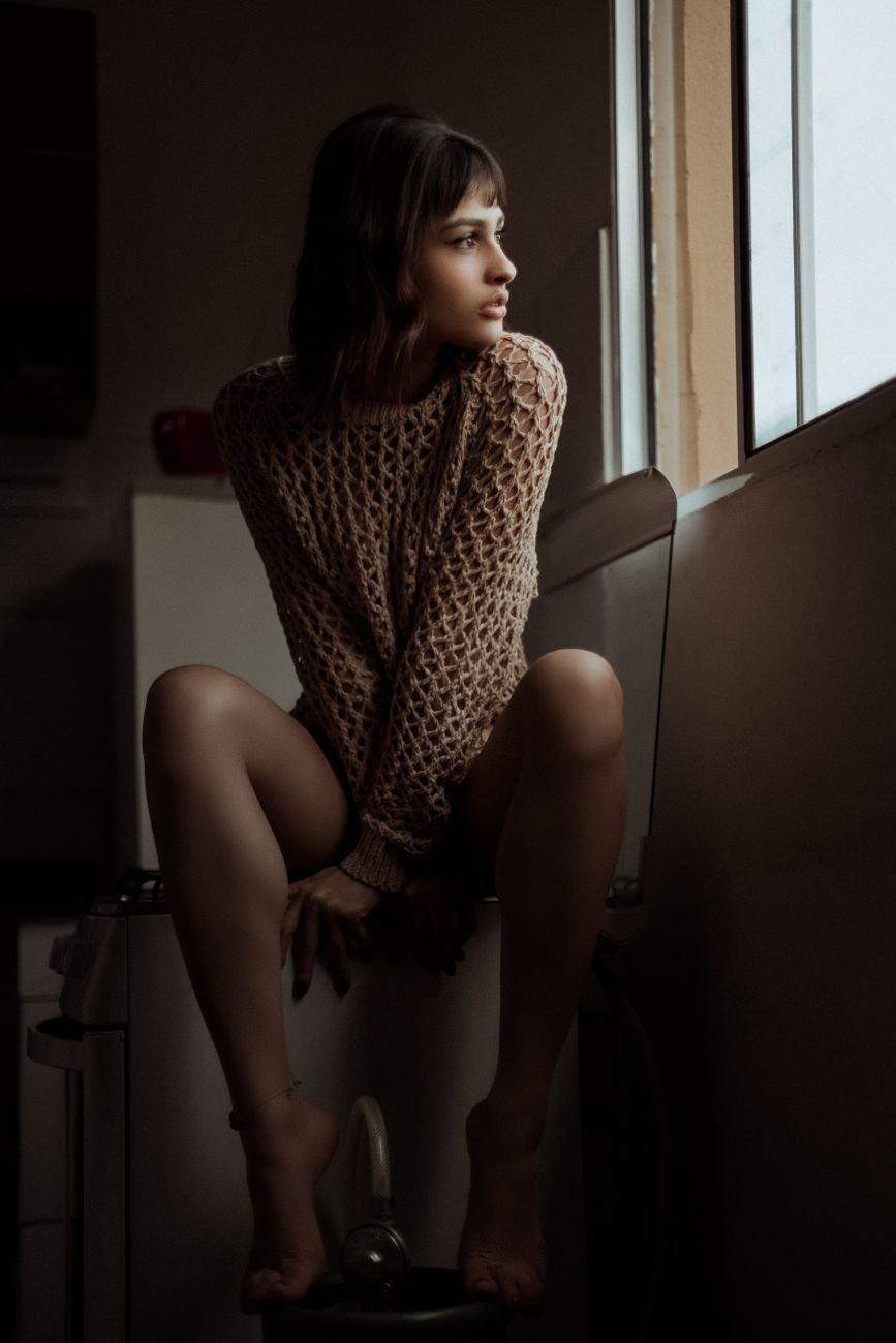 serious young ethnic female sitting near window in kitchen