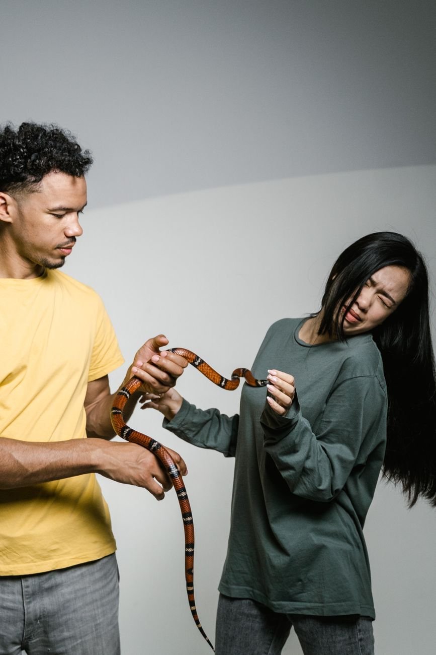 a fearful woman having ophidiophobia while standing next to a man who s holding a snake
